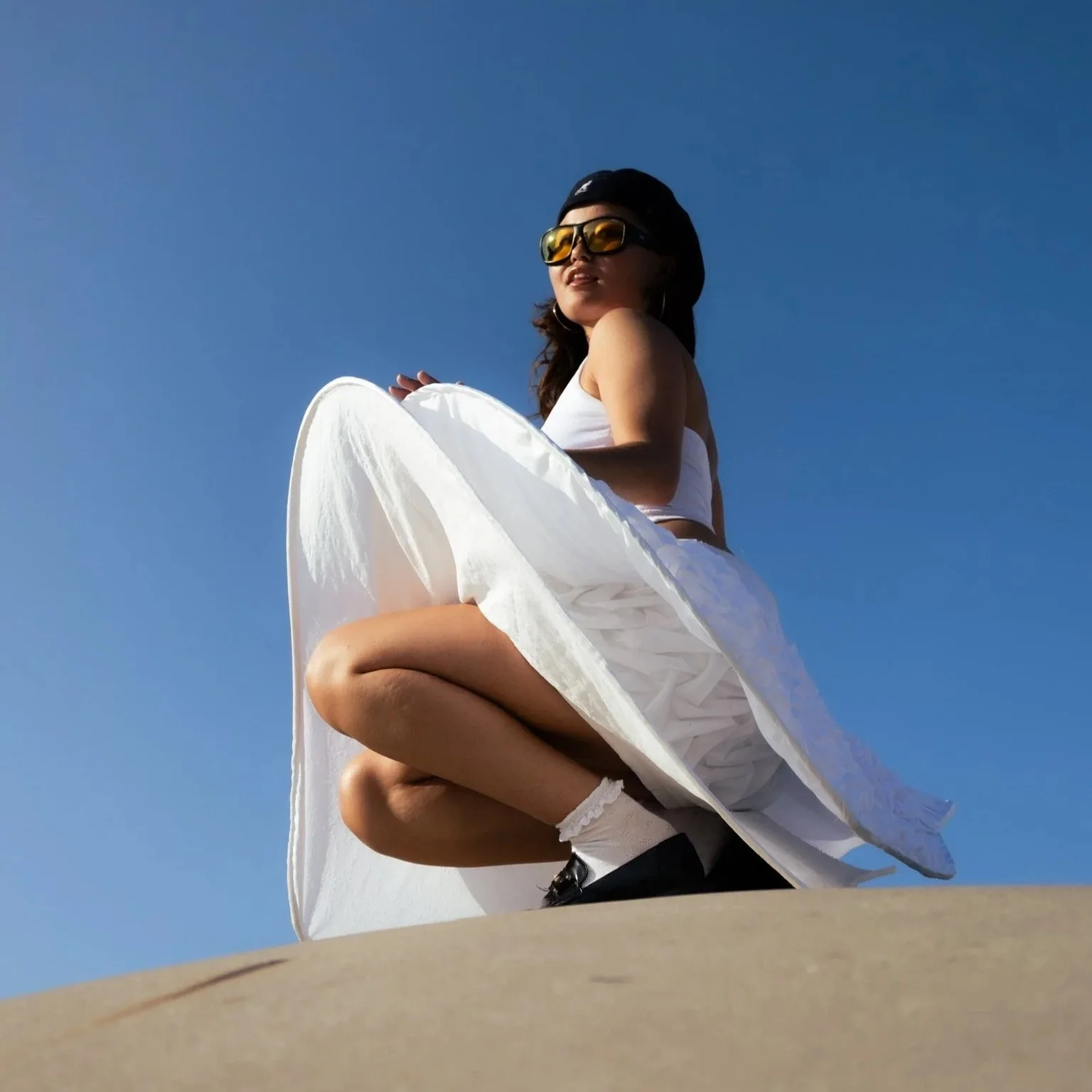 A woman squatting on a skateboard, wearing sunglasses, a black cap, a white crop top, white skirt, and white socks, against a clear blue sky.