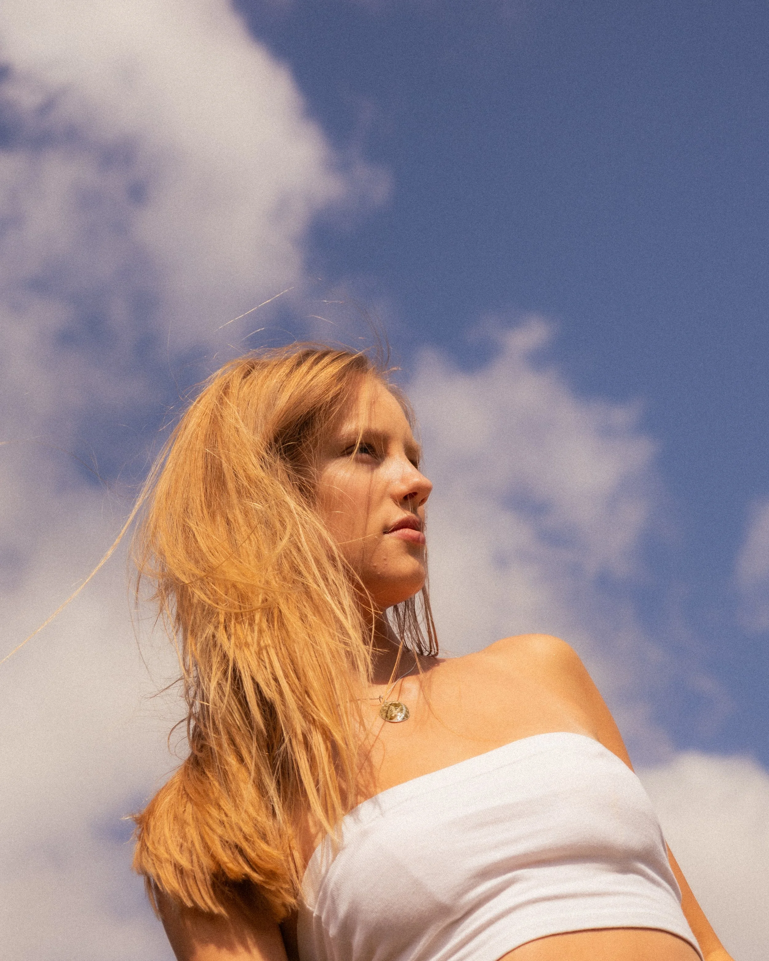 A young woman with long, wavy, blonde hair looking into the distance against a partly cloudy sky.
