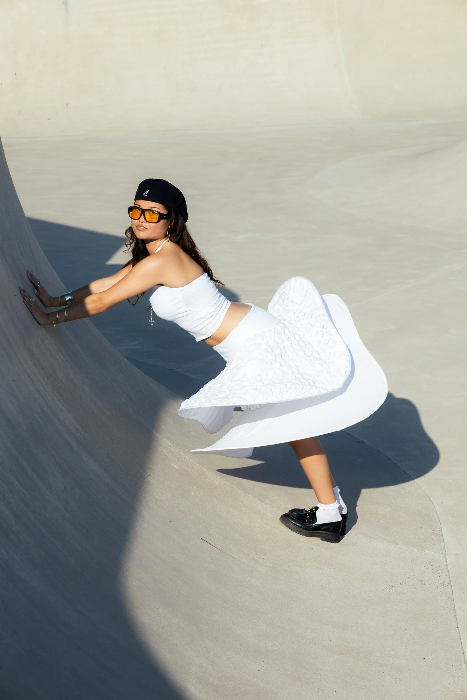 A woman wearing a white crop top, a white textured skirt, black shoes, white socks, a black hat, and orange sunglasses pushes against a sloped concrete surface at a skate park.