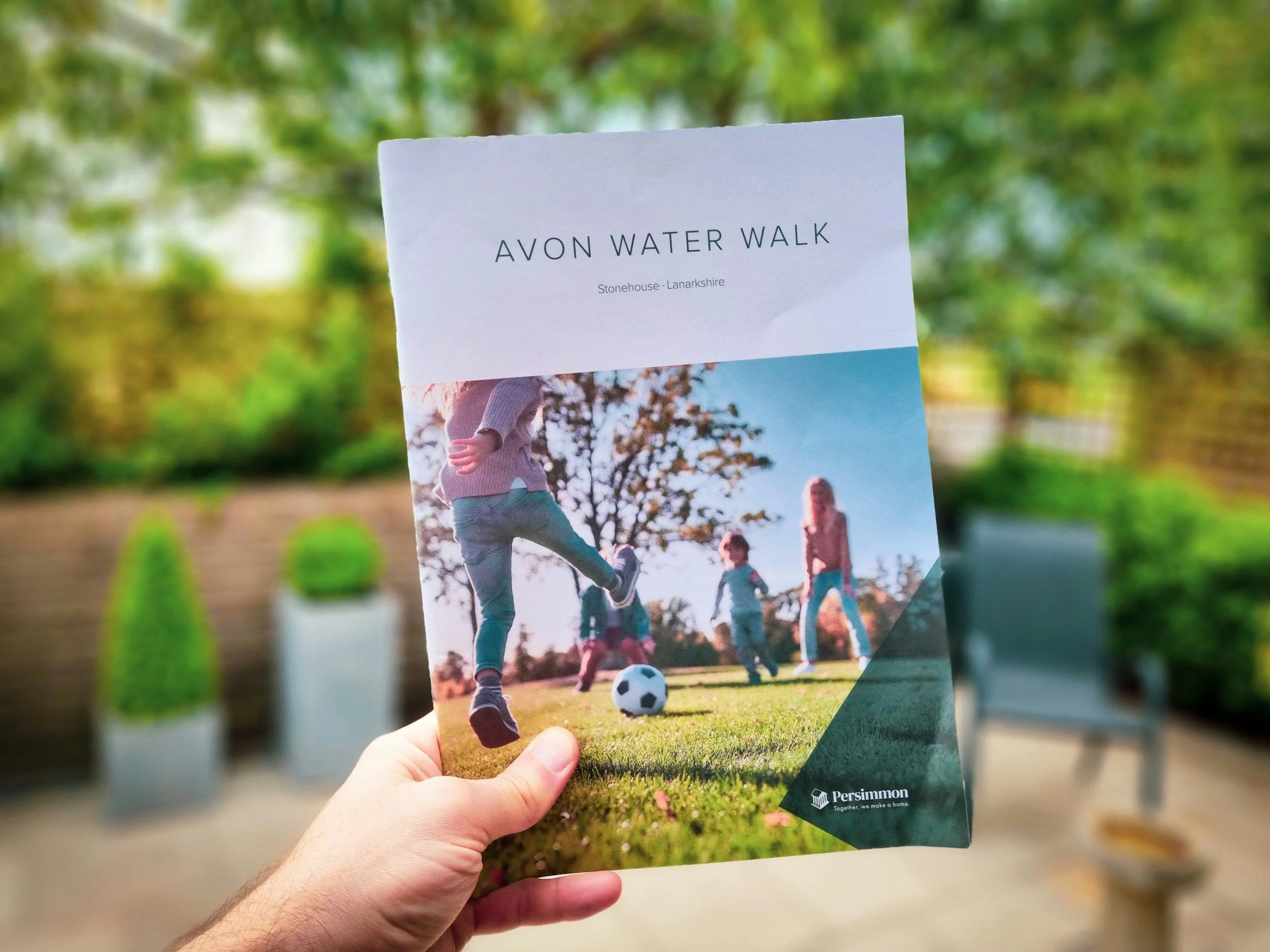 Hand holding a pamphlet titled 'Avon Water Walk' in a park with trees and benches in the background.