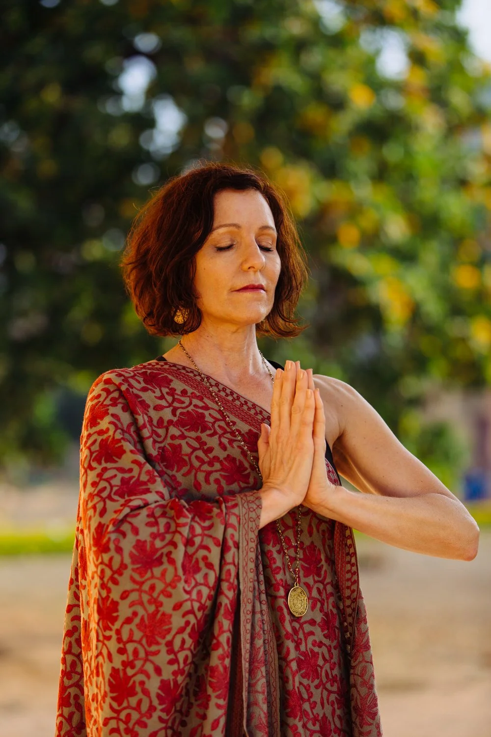 Lisa Bertke with closed eyes is standing outdoors with hands pressed together in a prayer position, wearing a patterned red and beige dress with a necklace. The background features green trees and natural sunlight.