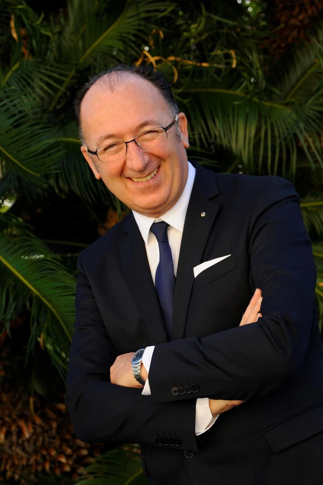 Uomo elegante sorride con braccia incrociate, indossa un completo nero e occhiali, sfondo di piante verdi tropicali.