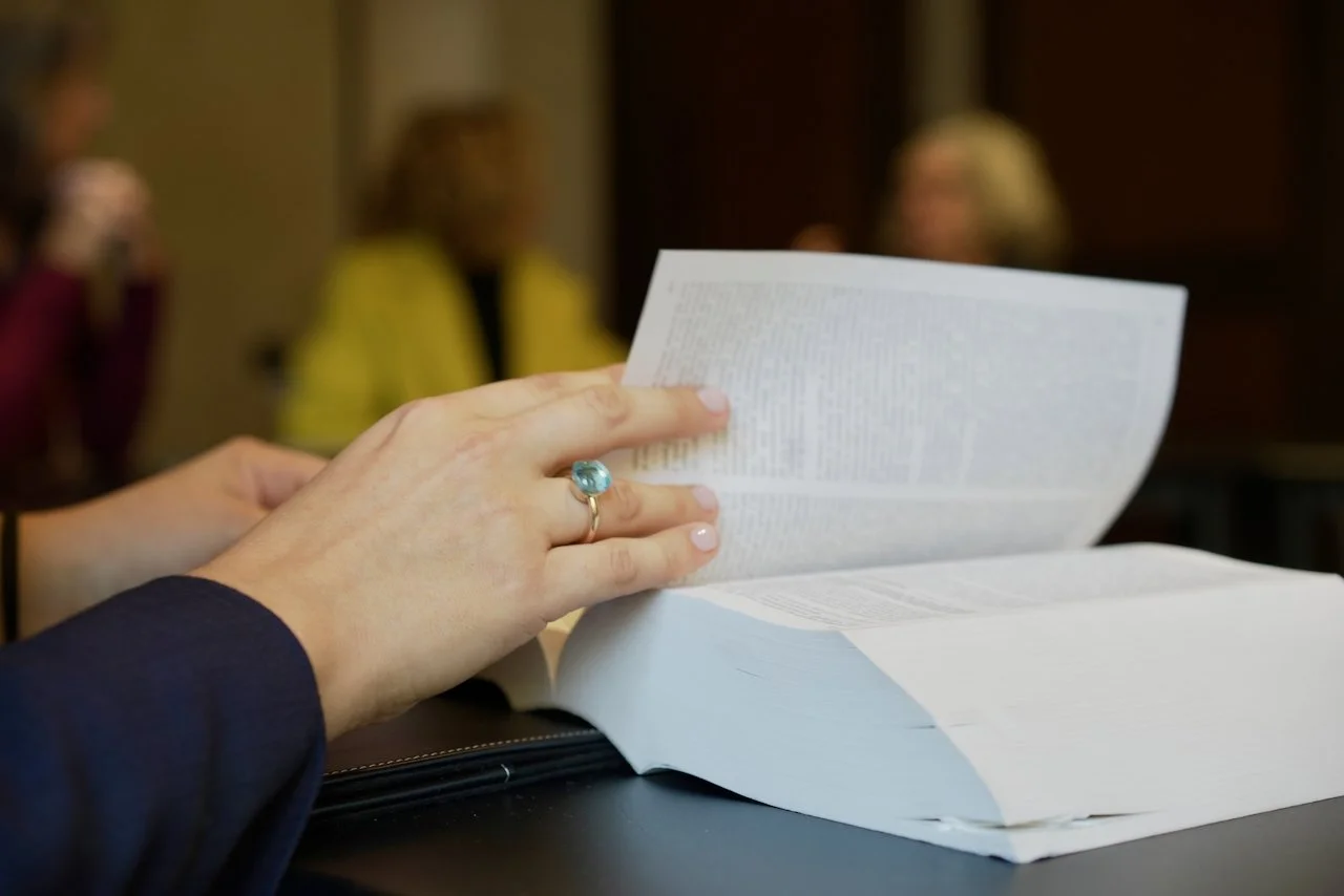 Una mano con un anello in pietra azzurra sta sfogliando un documento di grandi dimensioni su un tavolo, con persone sedute sullo sfondo in un ambiente interno.