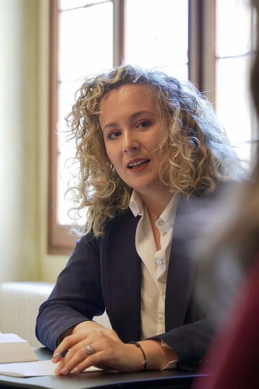 Donna con riccioli biondi e abbigliamento formale seduta a tavolo, sembra parlare o ascoltare in un ambiente illuminato dalla luce naturale di una finestra alle spalle.