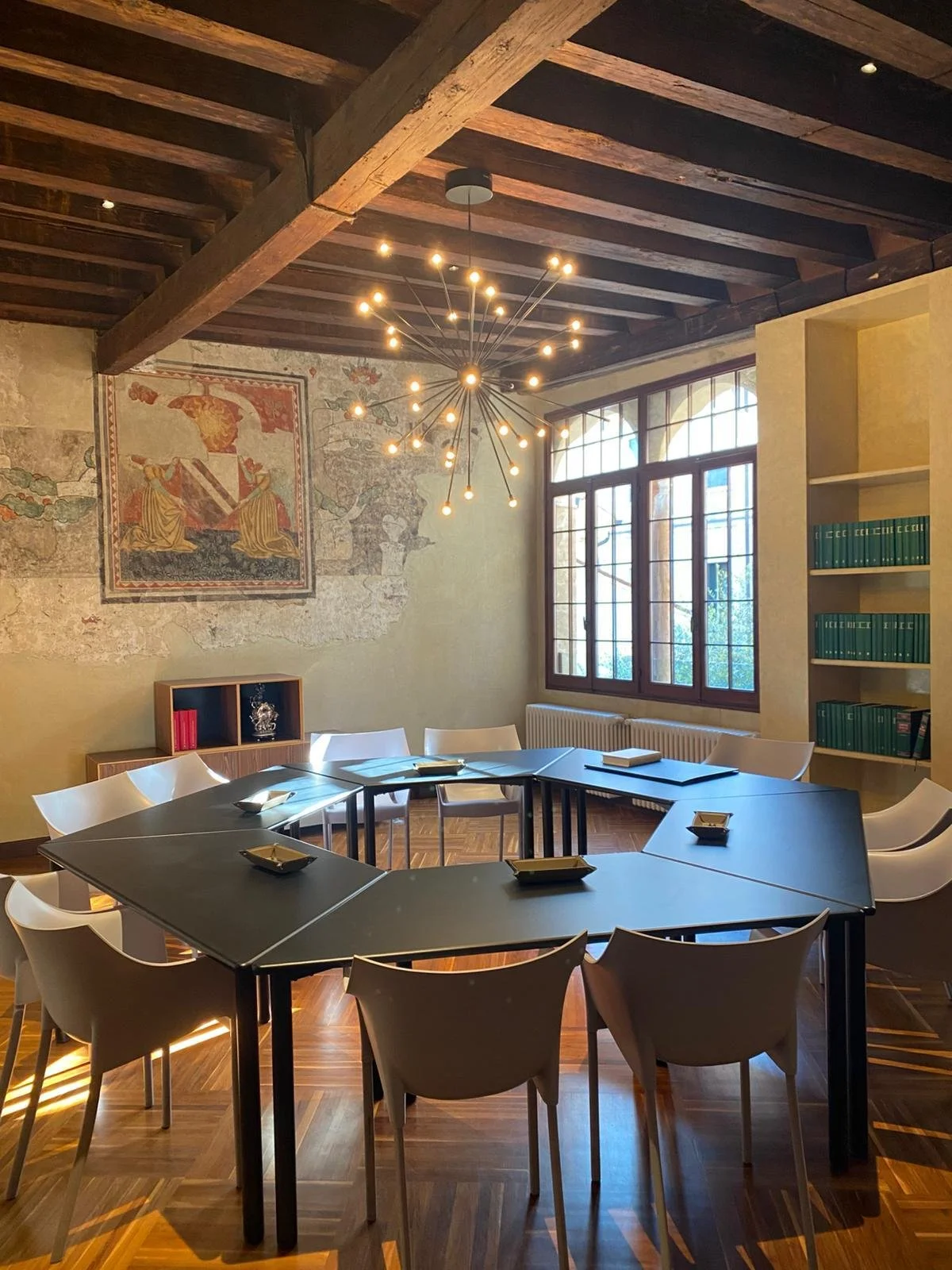 Sala riunioni con tavolo a forma di quadrato rotante, sedie bianche, grandi finestre, lampadario moderno e pareti decorate con affreschi.