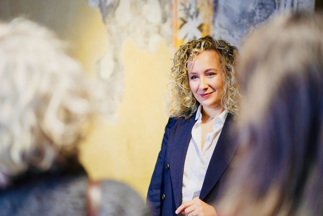 Donna con capelli riccioli chiari sorride mentre parla con un gruppo di persone, sfondo di parete decorativa.