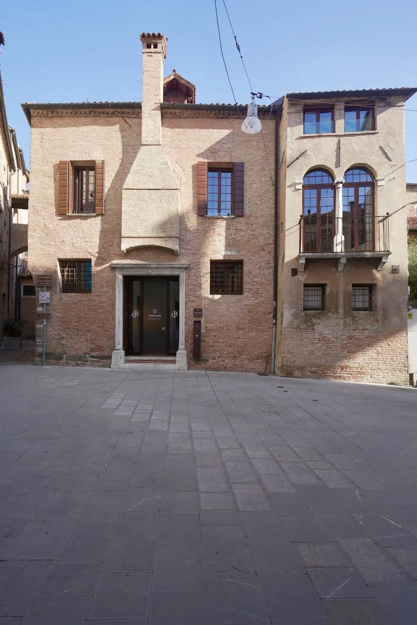 Edificio storico in pietra con due piani, finestre con tapparelle di legno, una porta d'ingresso incorniciata da colonne e una piccola balcony con ringhiera. La strada di fronte è pavimentata in pietra.