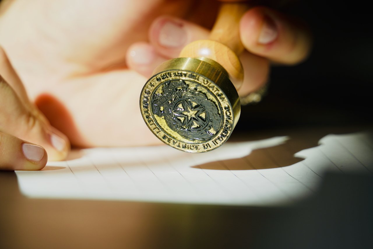 Una mano tiene un sello con un emblema circular, sostenuto sopra delle righe di carta, con il sole al centro e una stella a cinque punte.