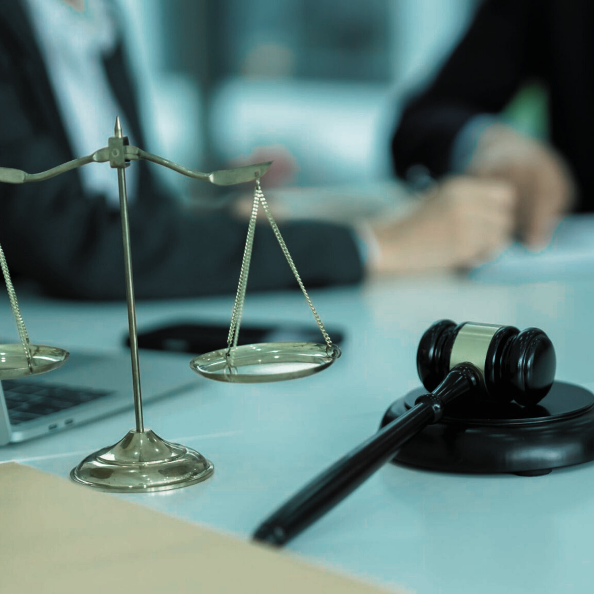 Scale model of justice scales, judge's gavel, and a person's hands in the background on a judicial desk.