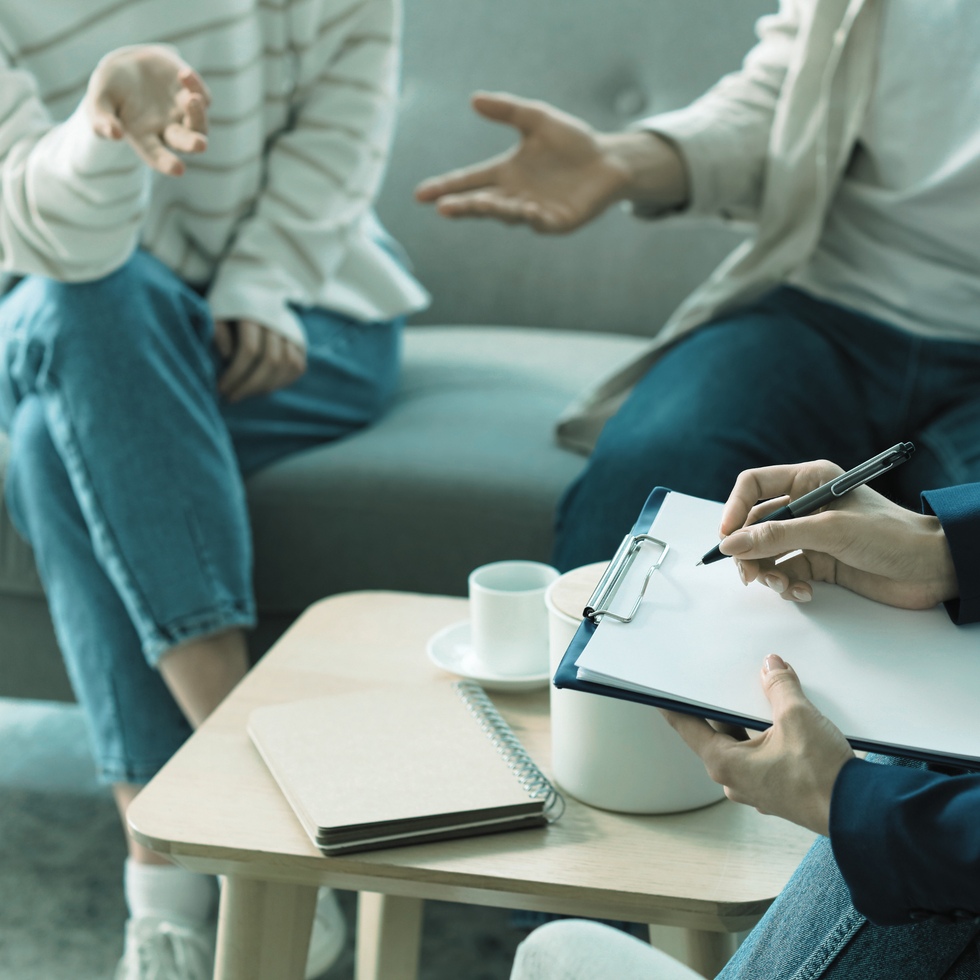 A therapist taking notes on a clipboard during a therapy session with a young woman sitting on a couch.