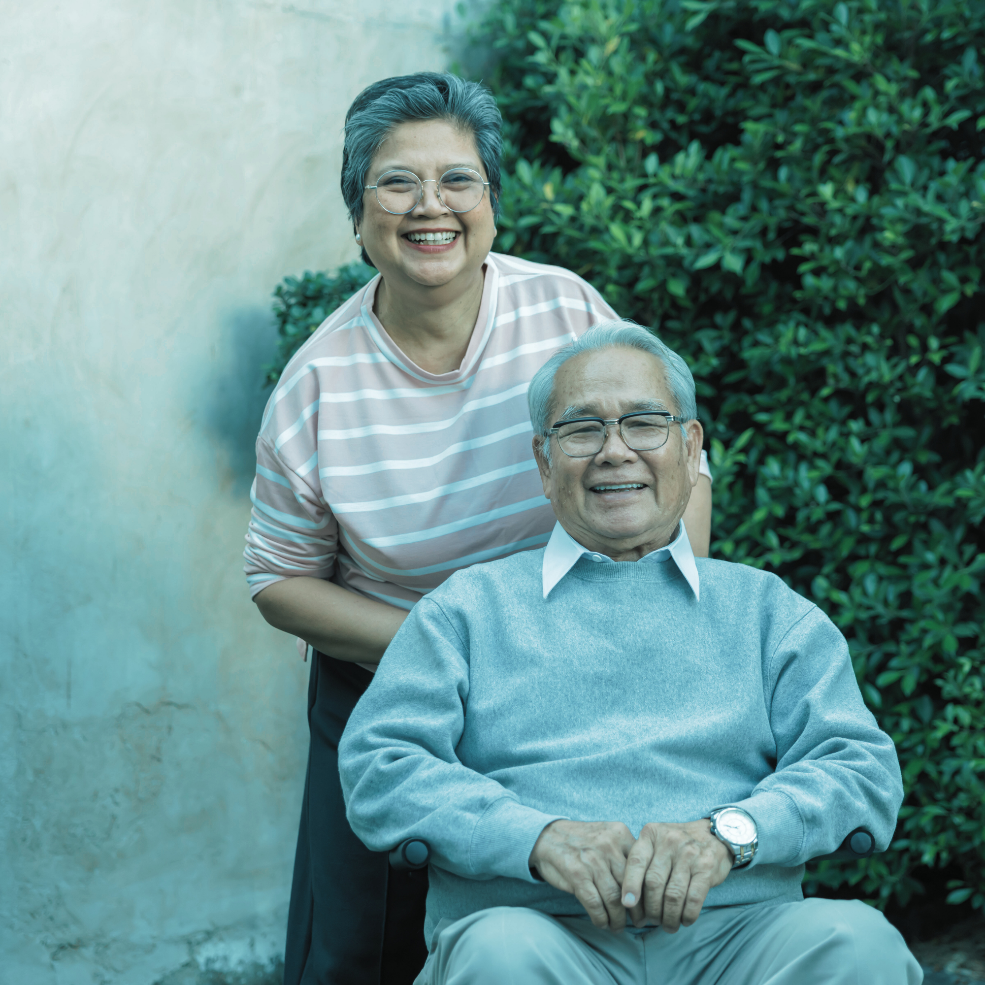 An elderly man sitting in a wheelchair outdoors, smiling, with a woman standing behind him, also smiling. They are near a wall and green bushes.