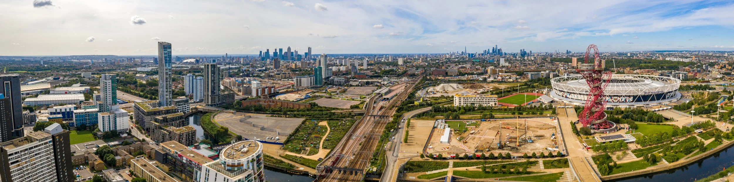 A panoramic view of a city skyline with many high-rise buildings, a stadium, and a twisting red and white tower, with visible roads, train tracks, and green spaces under a partly cloudy sky.
