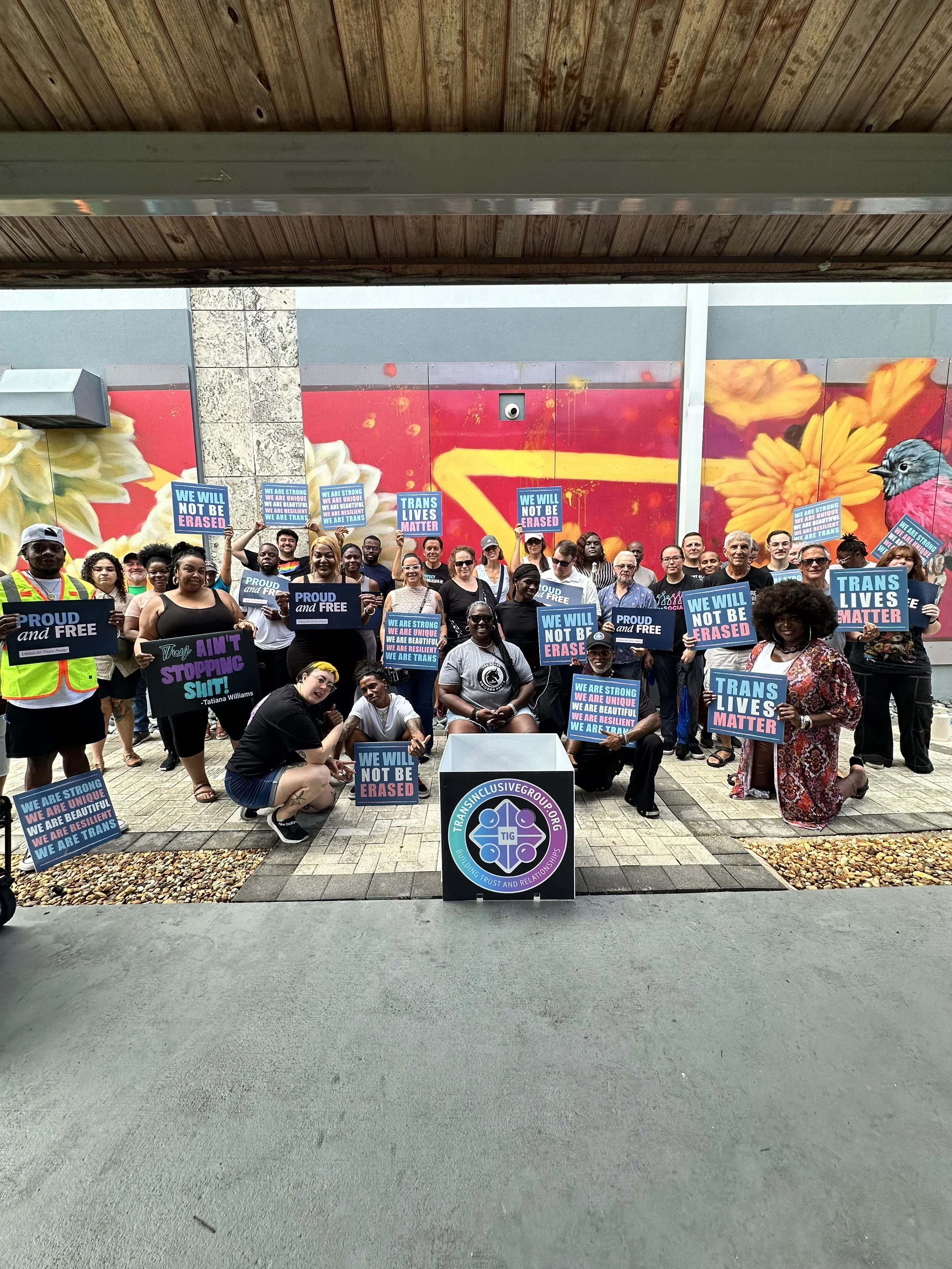 Group of diverse people gathered outdoors holding signs supporting trans rights and equality, with a colorful mural of flowers and a bird in the background.