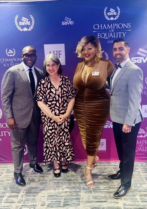 Four people posing in front of a backdrop that reads 'Champions of Equality Gala' with logos and laurels. They are dressed in formal attire, smiling and standing close together.