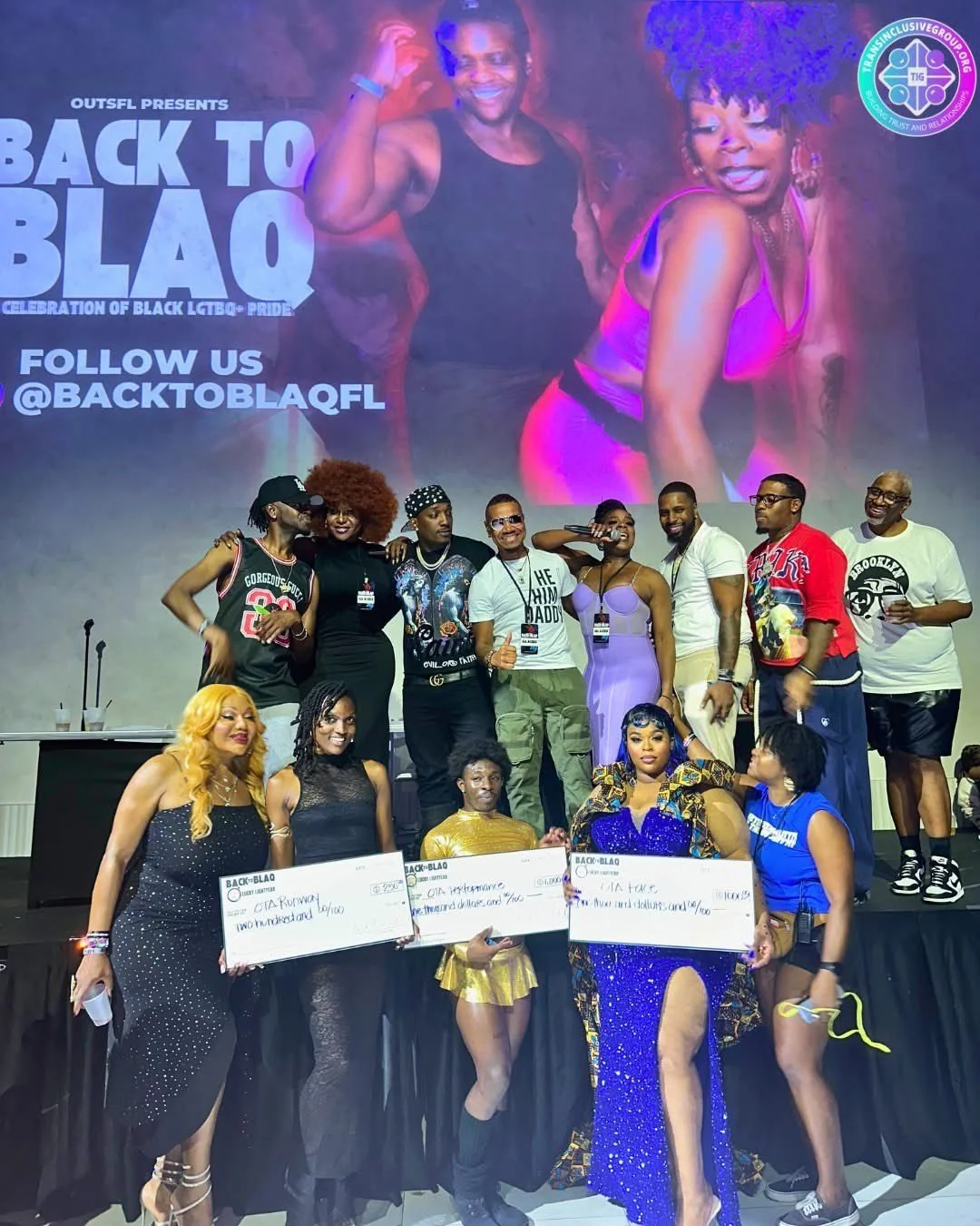 A group of people on stage celebrating at a Black LGBTQ+ pride event, with a large screen behind them displaying a photograph of two Black individuals in colorful outfits, and the text 'Back to Blaq' and 'Follow us @BackToBlaqFL'.