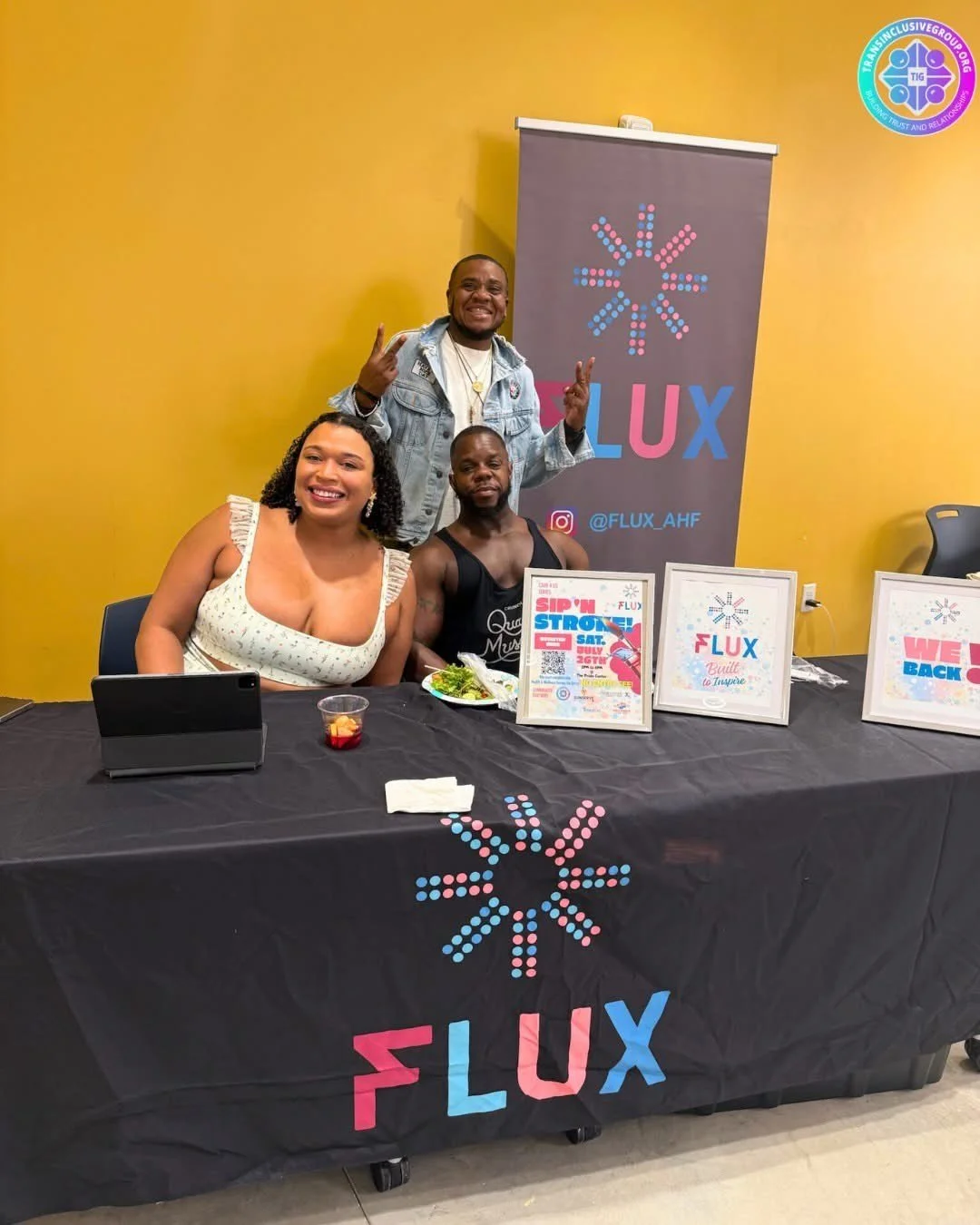 Three people sitting at a table with FLUX branding, two women and one man, with FLUX posters and a banner in the background, smiling and making peace signs, with a yellow wall behind them.