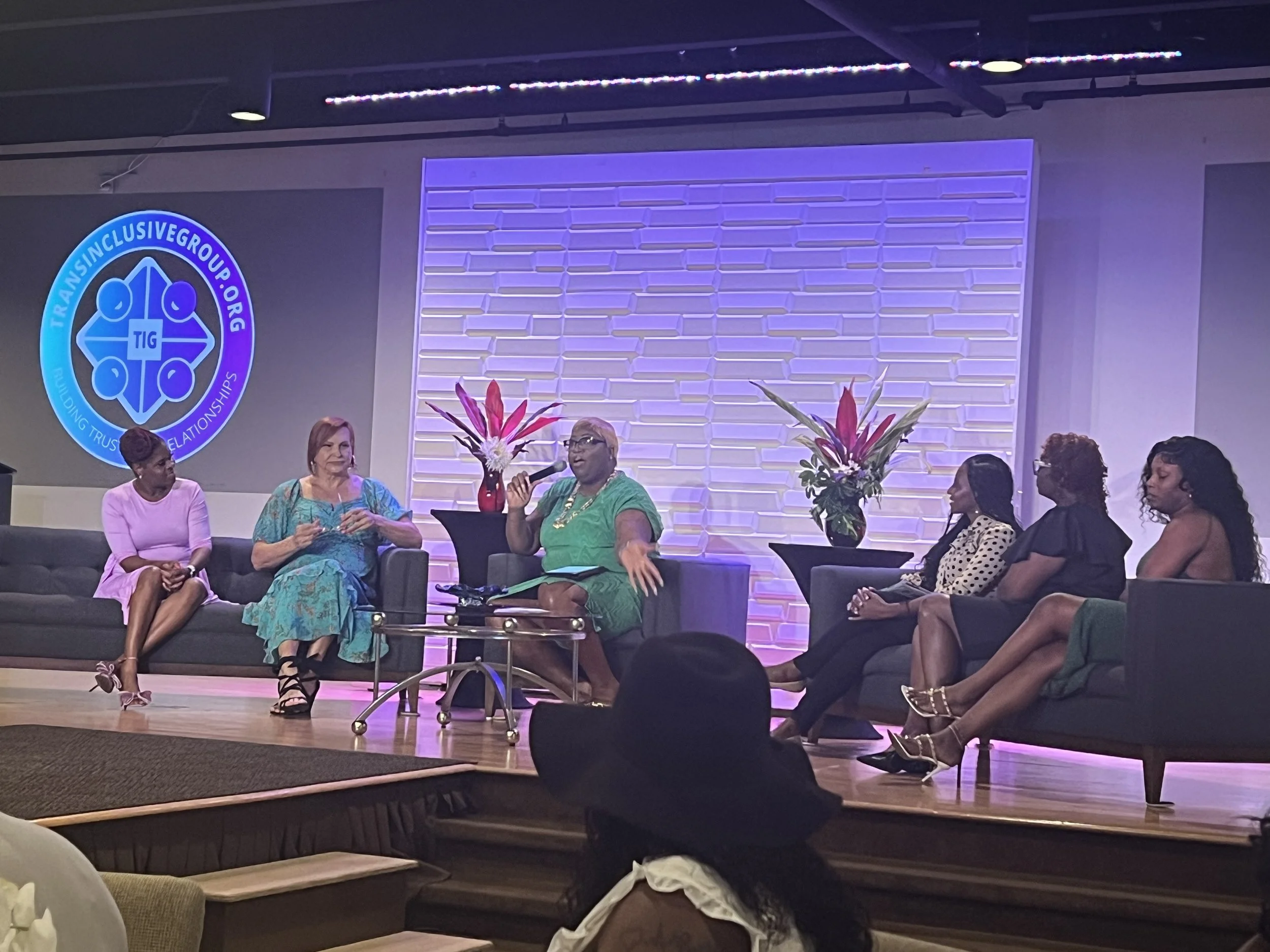 Panel discussion with six women sitting on stage, two with microphones. A screen with a logo reads 'TransInclusiveGroup.org'. Two large floral arrangements with pink and white flowers are on the stage.