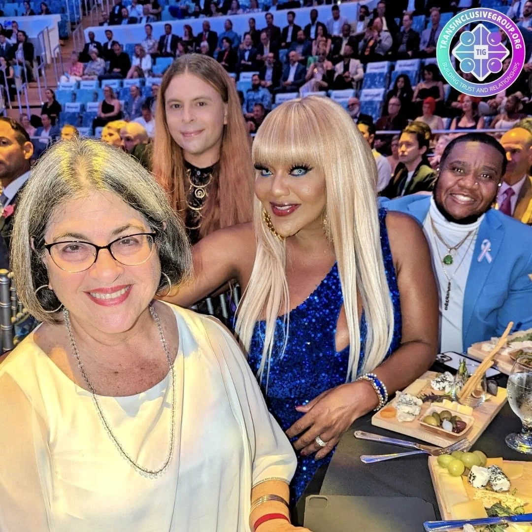 Four people sitting at a formal event with many attendees in the background. The woman in the foreground has glasses and gray hair, wearing a light-colored blouse and jewelry. Behind her, a blonde woman in a bright blue dress with sequins, bold makeu