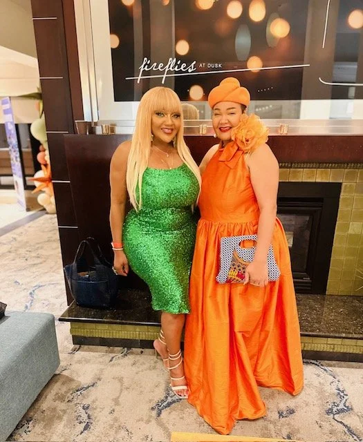 Two women are standing next to each other, smiling at a hotel or event venue lobby. One woman is wearing a bright green, knee-length, sequined dress with blonde hair and heels. The other woman is dressed in an orange, floor-length gown with a matchin
