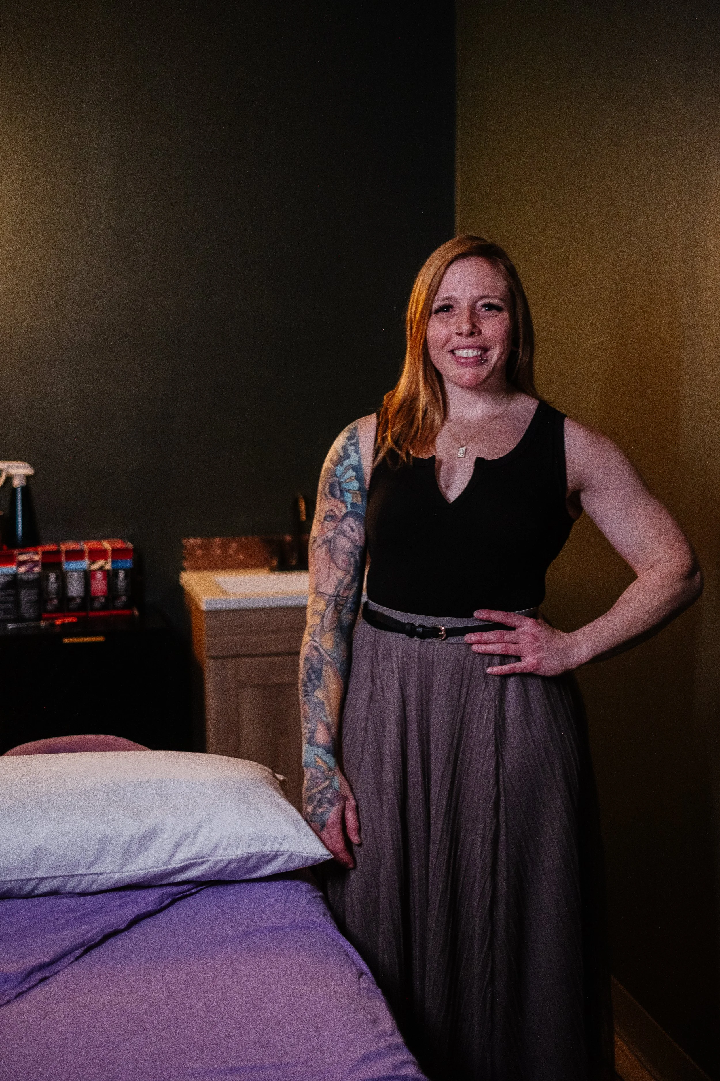 A woman with red hair and tattoos on her left arm standing in a bedroom, smiling and wearing a black sleeveless top and a long pleated skirt.