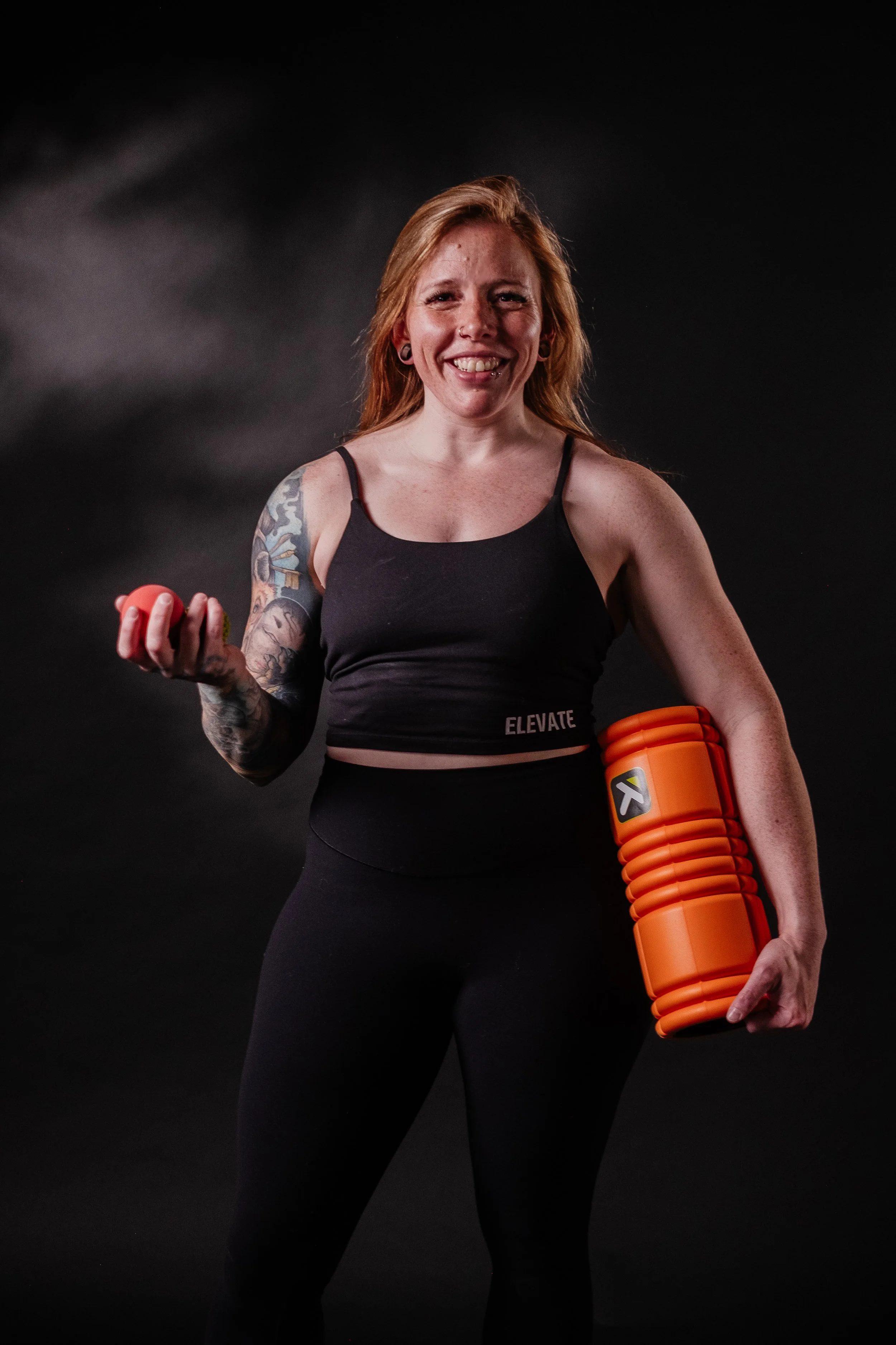 A smiling woman with tattoos on her left arm, wearing a black workout outfit, holding a small orange weight in her right hand and a foam roller in her left arm, standing in front of a dark background.