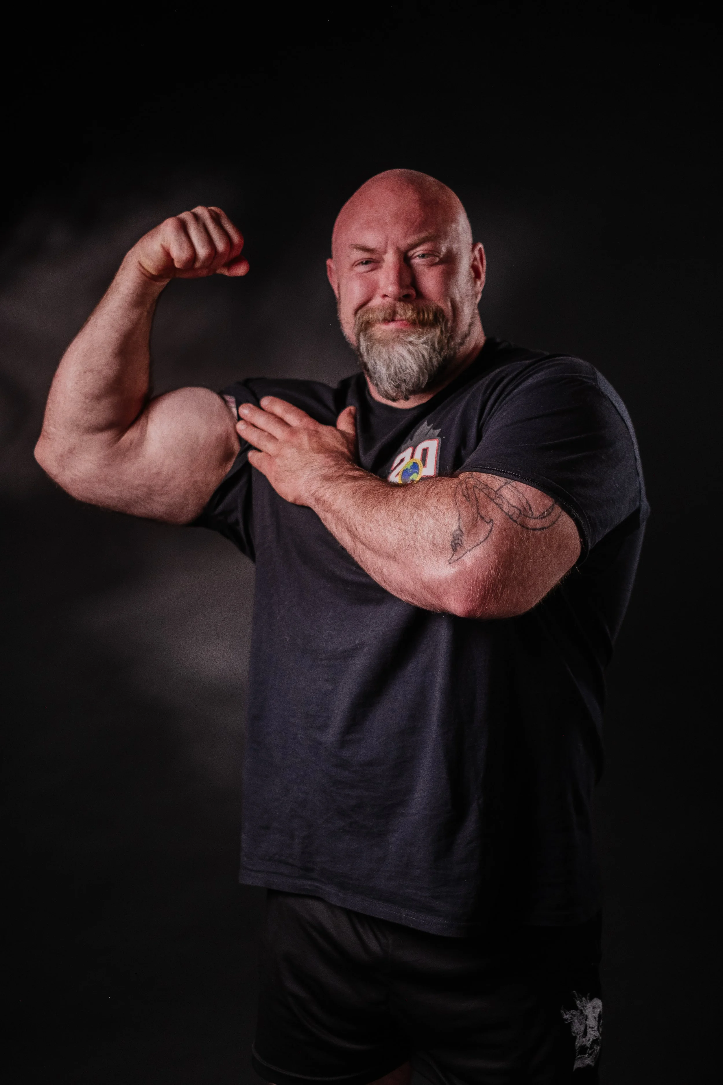 A bald man with a beard flexing his right arm, showing his bicep, while holding his left hand on his chest. He is wearing a black t-shirt and black shorts, and stands against a dark background.