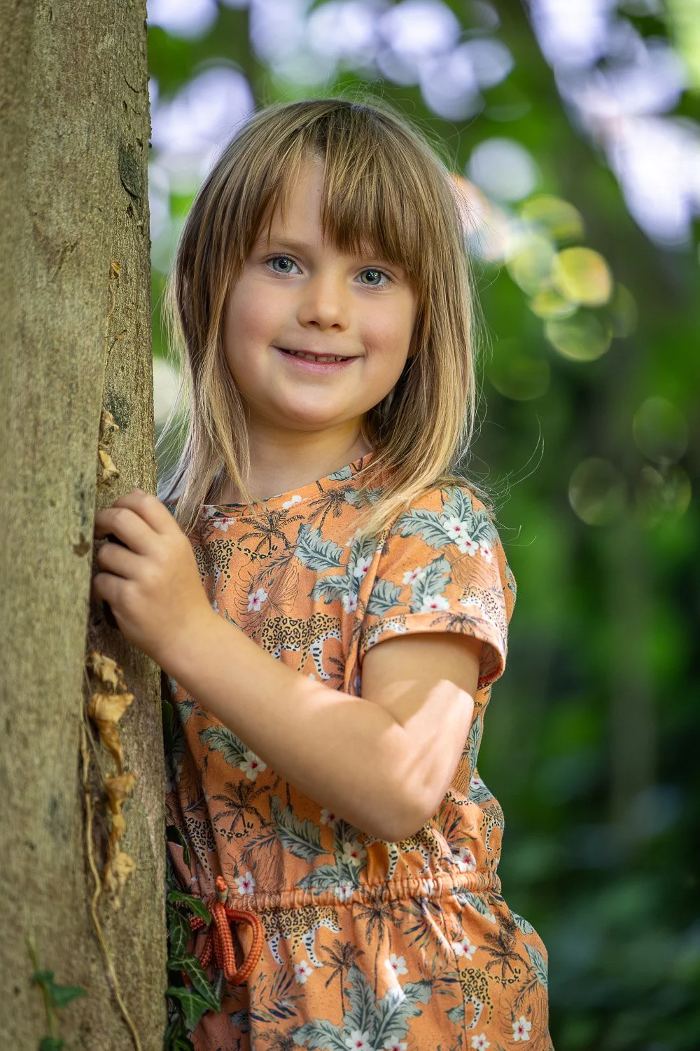 Ein lächelndes Mädchen mit braunen Haaren und blauen Augen, das im Wald hinter einem Baum hervorschaut.