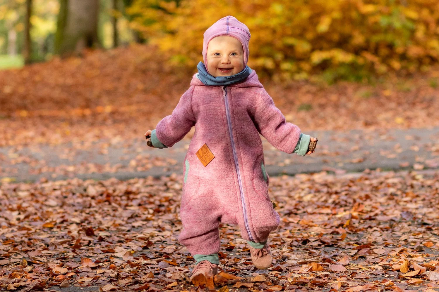 Ein kleines Kind in einem pinkfarbenen Overall, das im Herbst in einem Laub bedeckten Park lacht.