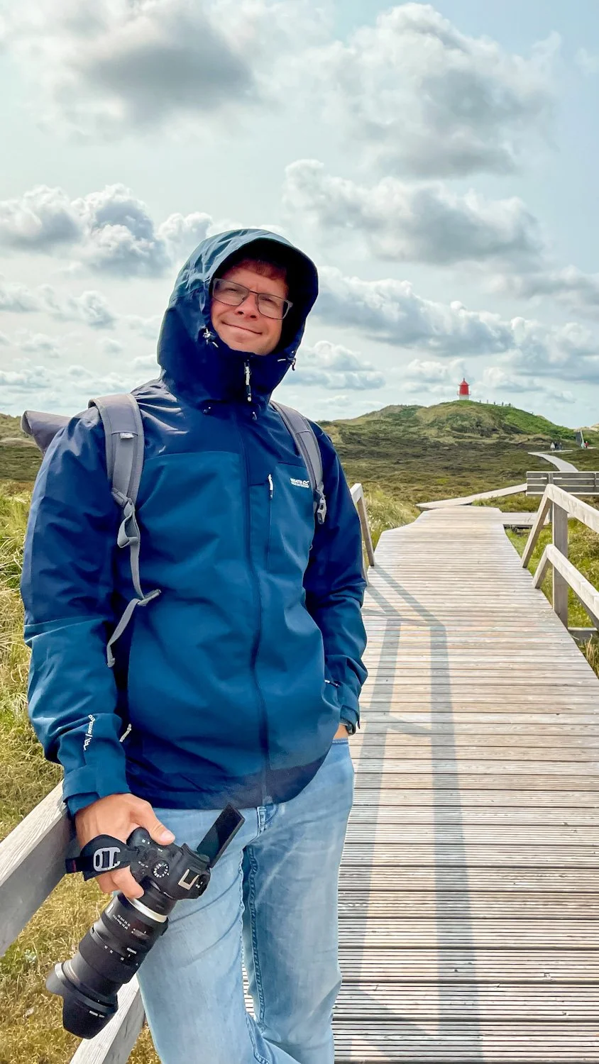 Junge Person in blauer Regenjacke mit Brille, Wanderschuhe und Rucksack steht auf einem Holzsteg in einer grünen, offenen Landschaft mit einem Leuchtturm im Hintergrund, bewölkter Himmel.