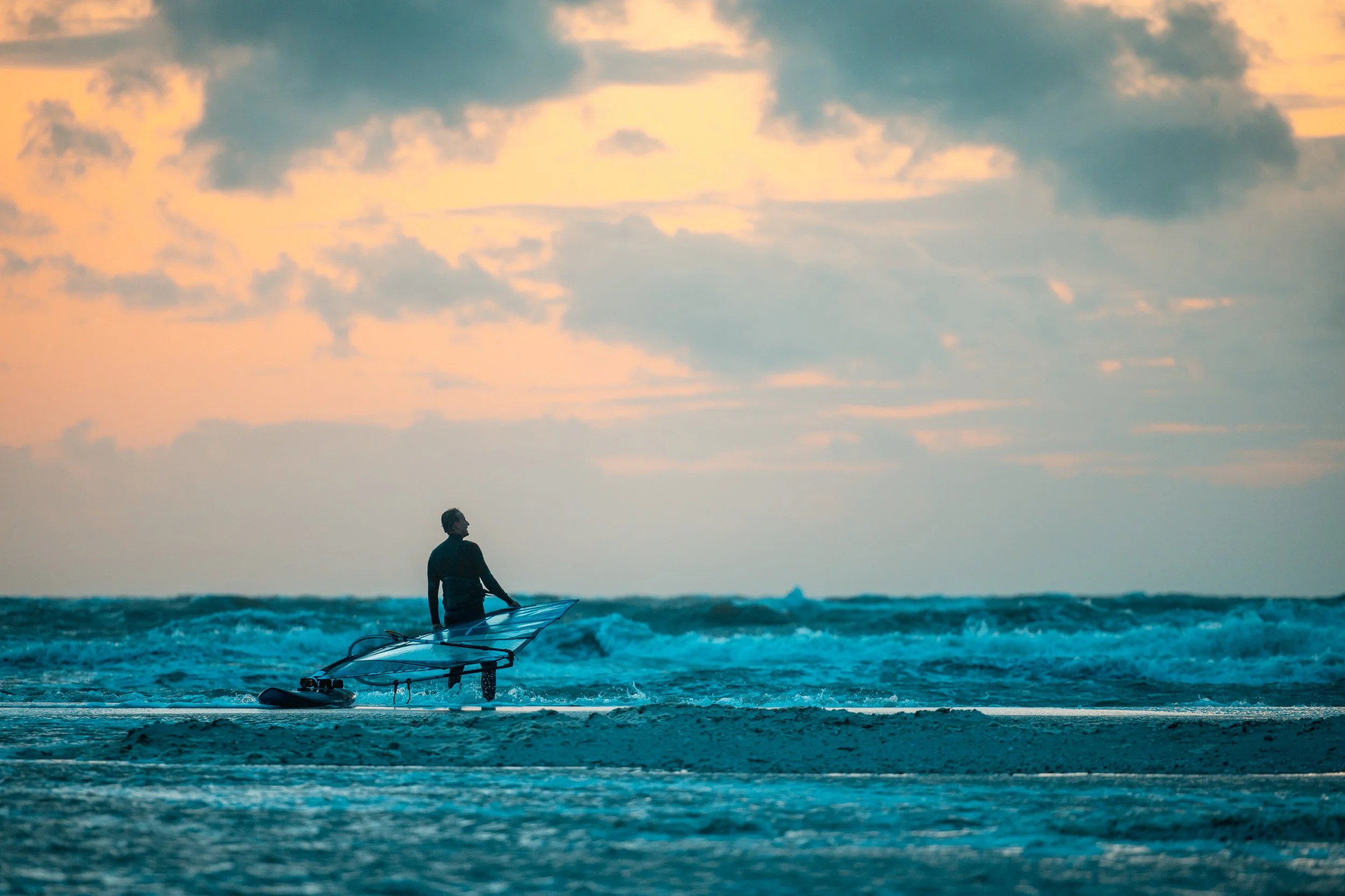 Ein Mensch steht mit einem Surfbrett im Wasser am Strand beim Sonnenuntergang.