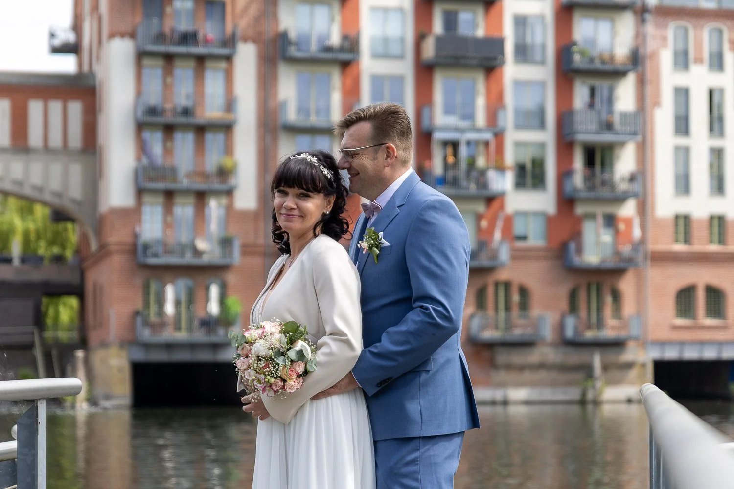 Ein glückliches Brautpaar bei der Hochzeit, stehend am Wasser, vor einem modernailen mehrstöckigen Wohngebäude in rotem Ziegelstein, mit Wasser und Balkon: Die Braut trägt ein weißes Kleid und hält einen Blumenstrauß, der Bräutigam trägt einen blauen