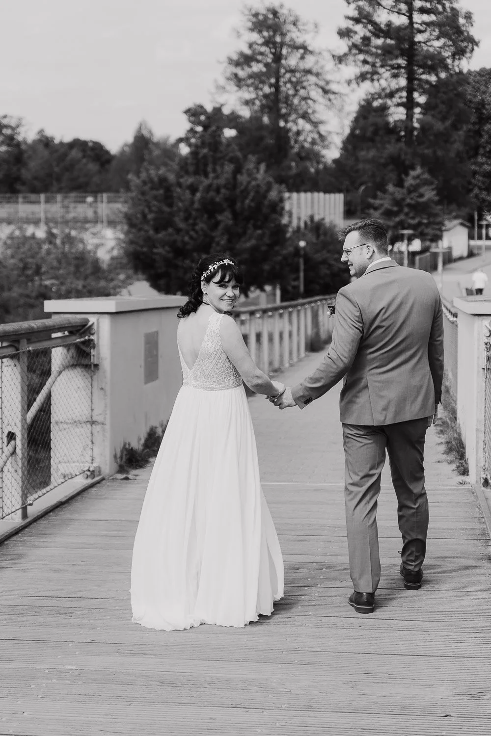 Ein Brautpaar, das sich auf einer Brücke in einem Park hält, in Schwarz-Weiß-Fotografie, lachend, im Anzug und Brautkleid.