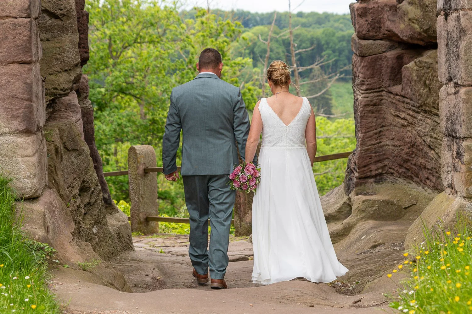 Ein Brautpaar geht Hand in Hand durch einen Torbogen in der Natur, die Braut trägt ein langes weißes Kleid und hält einen Blumenstrauß, die Szenerie ist grün und friedlich.