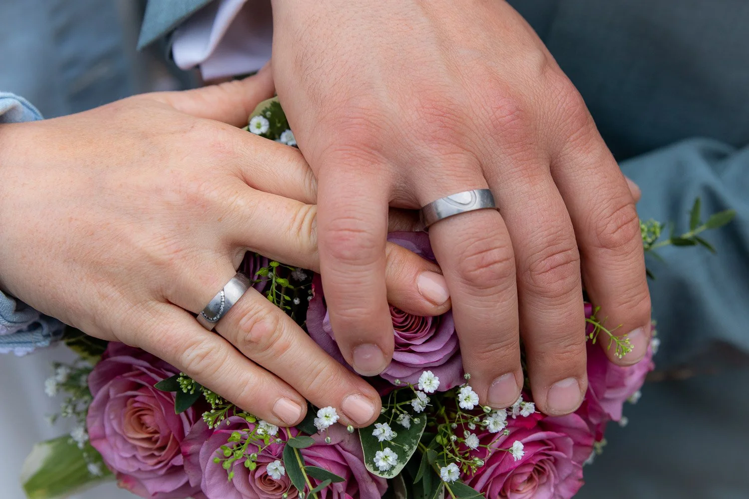 Zwei Hände mit Eheringen, die auf einem Blumenstrauß mit pinken Rosen und kleinen weißen Blumen ruhen.