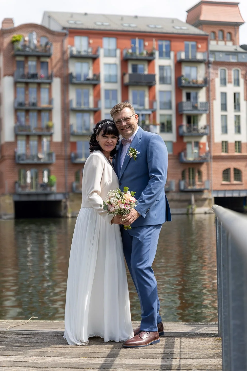 Ein frisch verheiratetes Paar, ein Mann in blauem Anzug und eine Frau in weißem Brautkleid, steht Hand in Hand auf einem Steg neben Wasser, vor einem roten Backsteingebäude mit Balkonen.