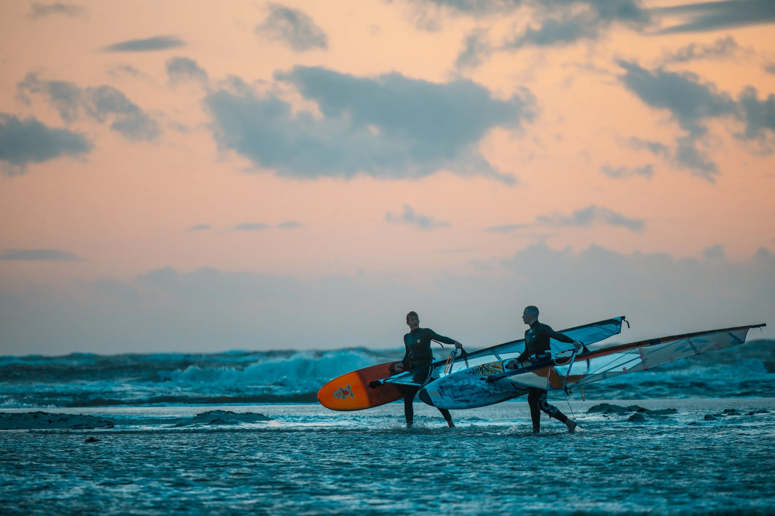 Zwei Windsurfer mit aufblasbaren Segeln am Strand bei Sonnenuntergang oder Sonnenaufgang, Wasser und Wolken im Hintergrund.