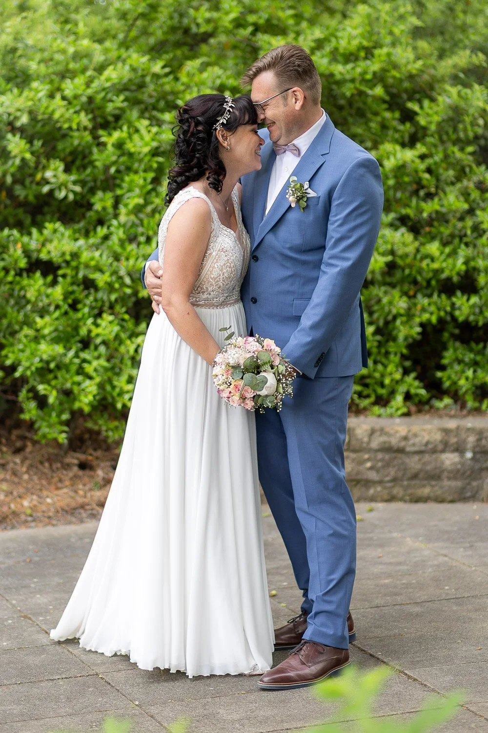 Ein brautpaar steht sich bei einer Hochzeit im Garten gegenüber, lächelt und hält sich an den Händen, während sie sich mit Stirn an Stirn berühren. Sie trägt ein weißes Brautkleid mit Spitze, er trägt einen blauen Anzug mit Fliege. Das Brautstrauß be