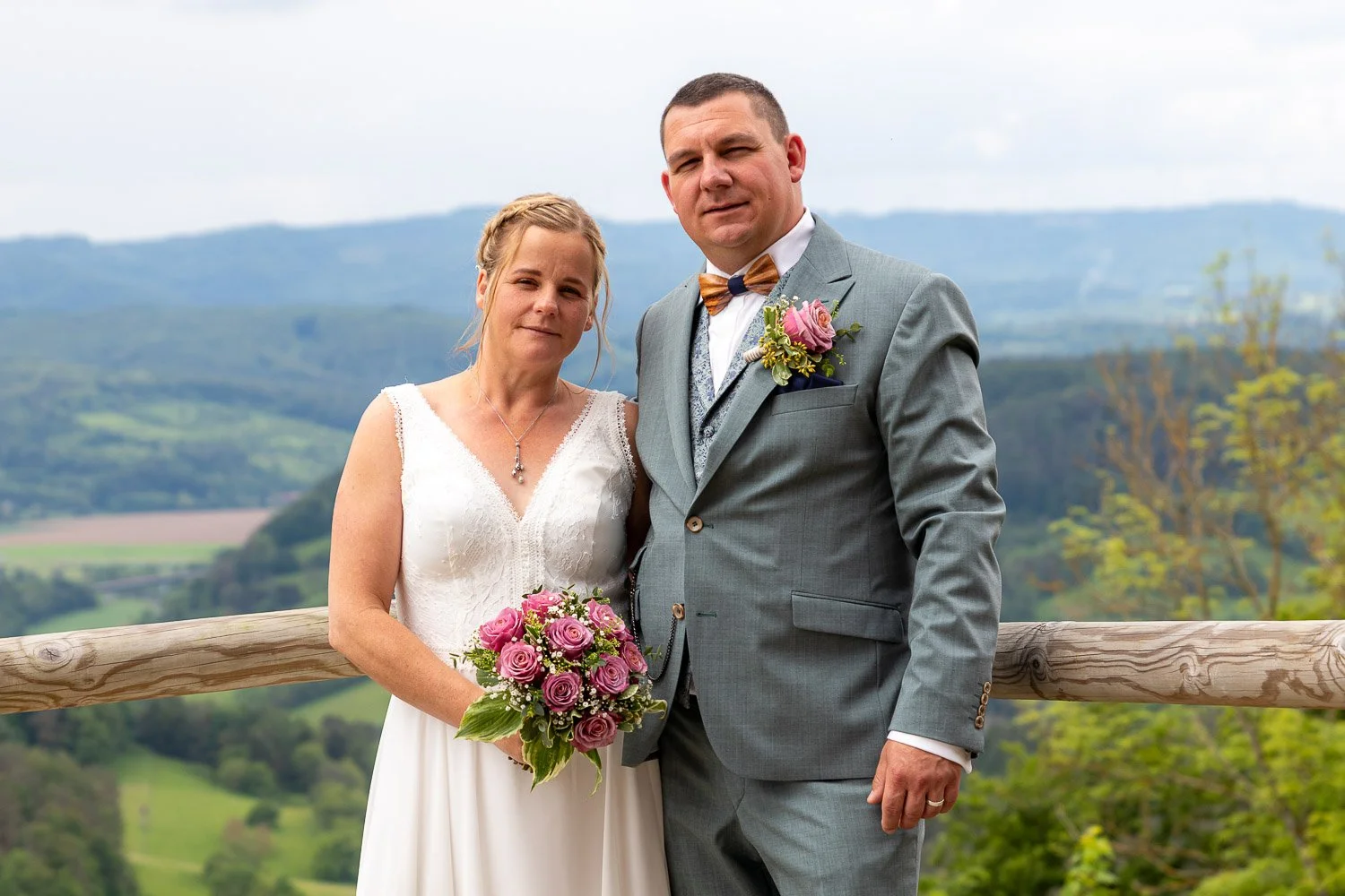 Braut und Bräutigam stehen an einem Holzgeländer in einer ländlichen Landschaft, die im Hintergrund Hügel und Bäume zeigt. Die Braut trägt ein weißes Kleid und hält einen rosa Blumenstrauß, während der Bräutigam einen grauen Anzug mit Fliege trägt.