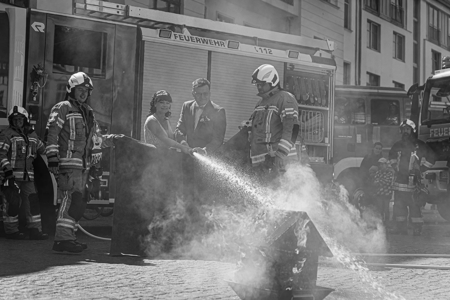 Feuerwehrleute löschen Brand bei einer Veranstaltung. Ein Mann und eine Frau, vermutlich Braut und Bräutigam, stehen zwischen den Feuerwehrleuten, die Wasser auf das Feuer sprühen. Auf der Rückseite stehen weitere Feuerwehrleute und Zuschauer.