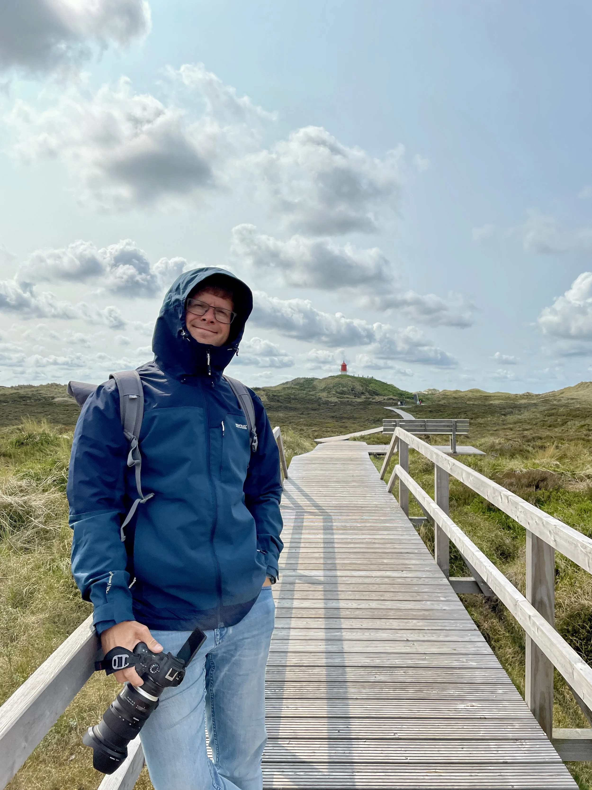 Junge Person steht auf einem Holzweg in einer ländlichen Umgebung, trägt eine blaue Jacke, Brille und hat einen Rucksack, hält eine Kamera in der Hand. Im Hintergrund sind grassbedeckte Hügel, eine Leuchtturm auf einem Hügel und ein bewölkter Himmel sichtbar.