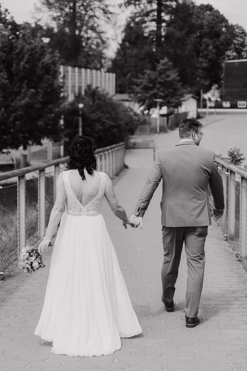 Braut in weißem Hochzeitkleid und Bräutigam in Anzug halten Händchen beim Spaziergang auf dem Bürgersteig.