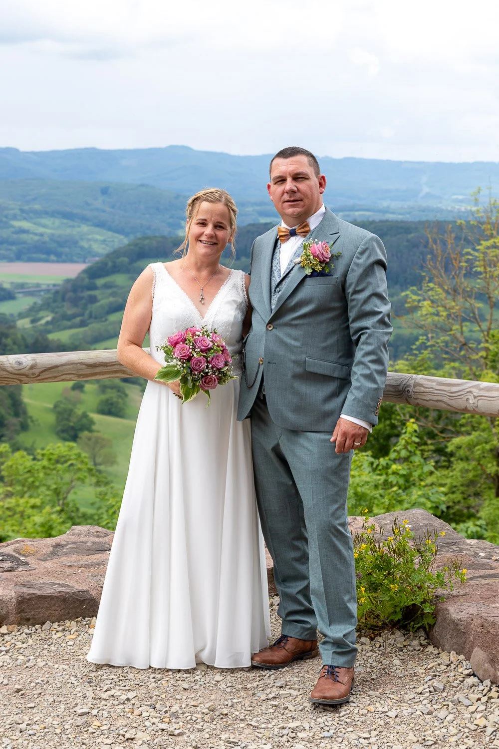 Ein Brautpaar steht auf einer Terrasse mit Blick auf grüne Hügel und Berge im Hintergrund. Die Braut trägt ein weißes Brautkleid und hält einen Blumenstrauß, während der Bräutigam einen grauen Anzug mit Krawatte und Blumenanstecker trägt.