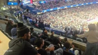 A crowd of people seated inside a sports arena during a game or event, with bright lights illuminating the court and audience.