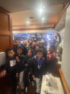 Group of children and teenagers gathered indoors, some smiling and waving at the camera, with a cityscape visible through the windows behind them.
