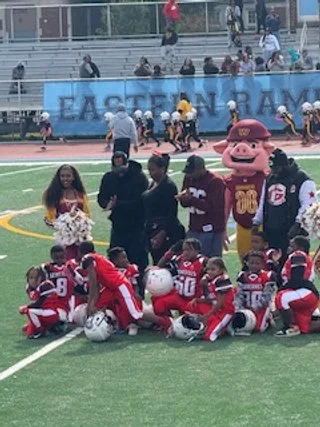 Youth football team in red and white uniforms with helmets, coaches, and a mascot in a maroon hoodie on a football field during a game or event.