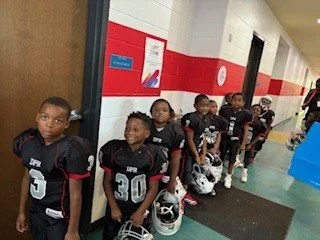 A group of young football players in black jerseys standing in a school hallway, holding helmets.