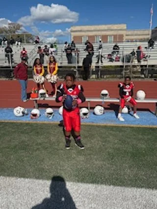 Football players in red uniforms on a field, with cheerleaders in yellow and red uniforms sitting on bleachers in the background, under a partly cloudy sky.