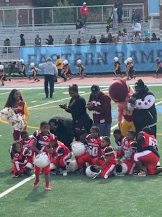 Young football players in red uniforms kneeling on the field after a game, with coaches and spectators nearby, and players in yellow running on the track in the background.