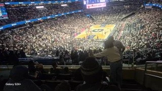 Crowd inside a large indoor sports arena, with a basketball court at the center, surrounded by spectators and some standing.