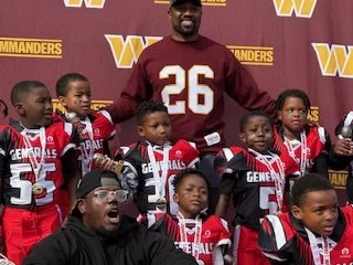 Group of young football players in red and black uniforms, with a coach or adult in a maroon shirt with the number 26, posing in front of a purple background with yellow and red text that says 'Commanders' and 'W'.
